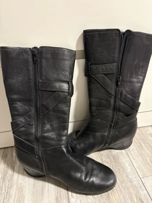 Svarta knähöga läderstövlar - Säljer ett par svarta knähöga stövlar i äkta läder med dragkedja på insidan och dekorativa sömmar. Stövlarna har en låg kilklack och stilren design som passar till många outfits. Perfekta för dig som vill ha både komfort och stil.