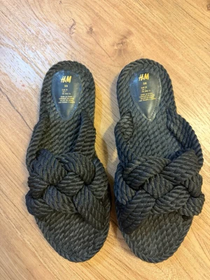 Svarta flätade sandaler från H&M - Säljer ett par svarta sandaler från H&M med grov flätad design i rep-liknande material. De har öppen tå och breda band över foten som ger en cool och trendig look. Perfekta för sommaren och riktigt bekväma att glida runt i.
