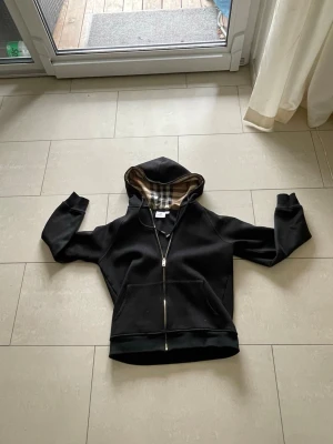 Svart hoodie från Burberry med dragkedja - Snygg svart hoodie från Burberry med dragkedja framtill och klassiskt rutigt foder i huvan. Hoodien har fickor på magen och ribbade muddar vid ärmslut och nederkant. Perfekt för dig som vill ha en stilren och exklusiv look.