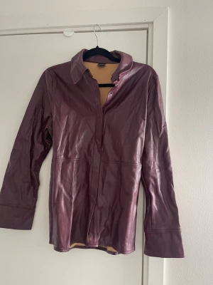 Vinröd skinnjacka med krage - Snygg vinröd skinnjacka med krage och tryckknappar vid ärmsluten. Jackan har en rak passform. Finns några revor i tyget vid axlarna och nacken, syns på bilderna.