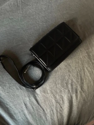 Svart quiltad axelväska - Snygg svart axelväska med quiltat mönster och lång justerbar rem. Väskan har magnetlås och stilren, rektangulär form. Perfekt för dig som gillar minimalistisk och trendig design. Materialet är konstläder som ger en lyxig känsla. Vet tyvärr inte var den är ifrån då jag köpte den på vinted💕💕