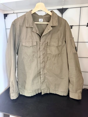 Olivgrön overshirt från C.P. Company - Stilren olivgrön overshirt från C.P. Company med två bröstfickor med lock och knapp, klassisk krage och knappstängning framtill. Tillverkad i lätt bomullsmaterial och har en avslappnad passform. Perfekt att bära över en t-shirt för en casual look. Dessutom köpt från Fashion stores leverantörer. 