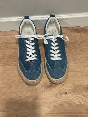 Blå denim sneakers med repdetalj - Snygga sneakers i blå denim med vita skosnören och en unik sula i repstil. Skorna har klassisk låg profil och vita detaljer runt snörningen. Har bara används en gång. Säljer pågrund av att de inte är min storlek. 
