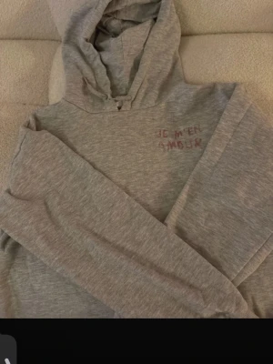 Grå hoodie med broderi - Grå hoodie med huva och dragsko, tillverkad i mjukt bomullsmaterial. På bröstet finns ett broderat textmotiv i lila rött som säger 'JE M'EN AMOUR'. Hoodien har långa ärmar och en avslappnad passform, perfekt för en chill och stilren look. Rätt så nopprig men det kan man säkert fixa med en noppermaskin 