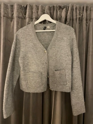Grå kofta från H&M med fickor - Mysig grå kofta från H&M med v-ringning och knappar framtill. Koftan har två ribbstickade fickor och ribbade muddar vid ärmslut och nederkant. Perfekt för lager på lager och enkel att matcha med olika outfits.