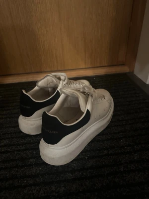 Vita Alexander McQueen sneakers - Säljer ett par klassiska vita sneakers från Alexander McQueen med svart detalj på hälen och tjock sula. Skorna har perforerade sidor, breda vita skosnören och logga både på plösen och hälen. Tillverkade i läder för en clean och stilren look.