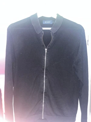 Svart ziptröja från Riley - Snygg svart ziptröja från Riley med klassisk krage och hel dragkedja framtill. Tröjan har lång ärm och är tillverkad i ett mjukt, tunt material som passar perfekt till lager på lager. Enkel och stilren design som funkar till det mesta.