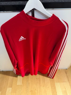 Röd Adidas sweatshirt med vita ränder - Säljer en röd sweatshirt från Adidas med klassiska vita ränder längs ärmarna och Adidas-logga på bröstet. Tröjan har rund halsringning och är tillverkad i ett mjukt, sportigt material som passar perfekt till träning eller chill.