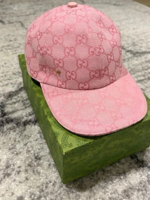 Rosa Gucci keps med GG-mönster - Snygg rosa keps från Gucci med ikoniskt GG-mönster över hela kepsen. Kepsen har en klassisk form och är tillverkad i ett tygmaterial som känns lyxigt och trendigt. Perfekt för dig som vill sticka ut och visa din stil. Använd ungefär 5 gånger