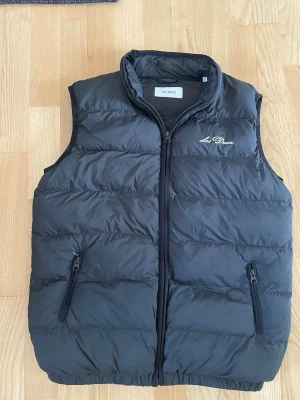 Svart dunväst från Les Deux - Snygg svart dunväst från Les Deux med broderad logga på bröstet. Västen har hög krage, två dragkedjeförsedda fickor och quiltad design. Perfekt för lager-på-lager och håller dig varm under kyliga dagar.