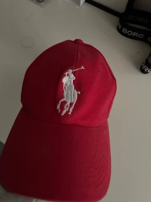 Röd keps från Polo Ralph Lauren - Snygg röd keps från Polo Ralph Lauren med den klassiska vita polospelaren broderad framtill och siffran 3 på sidan. Kepsen har böjd skärm och justerbar rem bak. Tillverkad i bomull för en bekväm och stilren look.