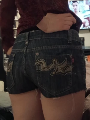Mörkblå jeansshorts med broderi - Säljer ett par mörkblå jeansshorts med råa kanter och snyggt broderade fickor bak. Klassisk femficksmodell med låg midja och slitna detaljer för en avslappnad look. Perfekta till sommaren!