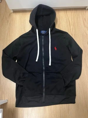 Svart hoodie från Polo Ralph Lauren - Snygg svart hoodie med dragkedja från Polo Ralph Lauren. Klassisk design med vit dragsko i huvan och röd broderad logga på bröstet. Hoodien är i fleece och har kängurufickor framtill. Perfekt för en chill och stilren look. Zipen är i lite dålig kvalite och lite plastig annars perfekt! 