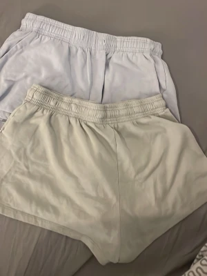 Två par ljusa shorts med resårmidja - Två par enkla shorts i ljusa färger, båda med elastisk midja och avslappnad passform. Perfekta för varma dagar och chill stunder. Materialet känns mjukt och lätt, vilket gör dem sköna att bära.