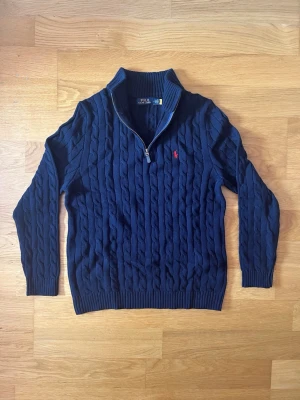 Mörkblå kabelstickad tröja Polo Ralph Lauren - Kabelstickad mörkblå tröja från Polo Ralph Lauren med halv dragkedja och ribbade muddar. Klassisk röd logga broderad på bröstet och hög krage. Perfekt för lager på lager och riktigt snygg till jeans.