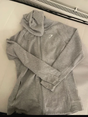 Ljusgrå hoodie från Gymshark - Snygg ljusgrå hoodie från Gymshark med dragkedja framtill och diskret logga på bröstet. Tillverkad i mjukt material och har en klassisk huva samt långa ärmar. Perfekt för en sportig och avslappnad stil.