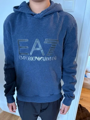 Blå hoodie från Emporio Armani EA7 - Snygg blå hoodie från Emporio Armani EA7 med stor logga i silver på bröstet. Klassisk huva och ribbade muddar vid ärmslut och nederkant. Perfekt för dig som gillar stilrena och trendiga plagg med exklusiv känsla.