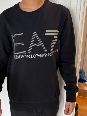 Svart sweatshirt från Emporio Armani - Snygg svart sweatshirt från Emporio Armani med stort EA7-tryck i vitt och grått på bröstet. Tröjan har rund halsringning, långa ärmar och ribbade muddar vid ärmslut och nederkant. Perfekt för en stilren och trendig look.