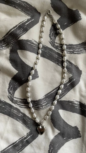 Pärlhalsband med hjärtberlock - Snyggt halsband med vita pärlor och en silverfärgad hjärtberlock. Halsbandet har metallspänne och justerbar kedja. Perfekt accessoar för att lyfta din outfit med en söt och trendig detalj.