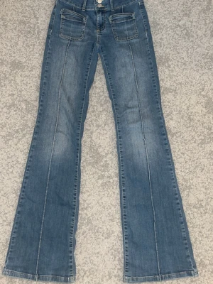 Blå bootcut jeans med fickor fram - Säljer ett par blå bootcut jeans med markerade sömmar och två stora fickor framtill. Jeansen har även klassiska bakfickor och stängs med knapp och dragkedja. 