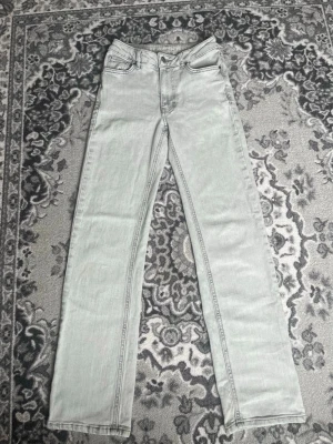 Ljusgrå raka jeans - Säljer ett par ljusgrå jeans med rak passform och klassisk femficksdesign. Jeansen har hög midja och är tillverkade i denim med synliga sömmar. Perfekta för en clean och stilren look.