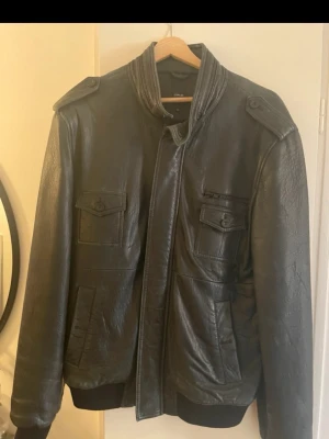 Svart skinnjacka med fickor - Cool svart skinnjacka med hög krage och knäppning framtill. Jackan har två bröstfickor med lock och knappar samt två sidofickor. Ribbad mudd vid ärmslut och nederkant ger en snygg passform. Perfekt för dig som vill ha en stilren och tidlös look. Från märket Emilio xl men sitter som L