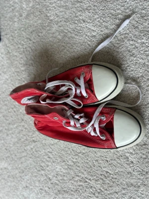Röda höga sneakers från Converse - Säljer ett par klassiska röda Converse sneakers med högt skaft och vita gummidetaljer. Skorna har vita snören och tåhätta, samt silverfärgade öljetter. Perfekt för dig som vill ha en ikonisk och cool stil. Passar ca 38-39❤️