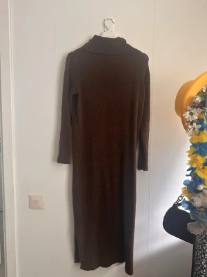 Brun stickad poloklänning Pieces - Mysig brun lång stickad klänning från Pieces med polokrage och långa ärmar. Perfekt för kyligare dagar och enkel att styla med boots eller sneakers. Klänningen har en rak passform och mjukt material.