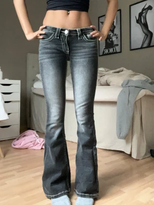 Grå lågmidjade bootcut jeans - Säljer ett par grå bootcut jeans med tydliga vita kontrastsömmar och slitningar framtill. Jeansen har låg midja, fem fickor och markerade bakfickor med dekorativa sömmar. Perfekta för en trendig och avslappnad look.