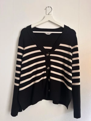 Svartvit randig kofta från Bubbleroom - Snygg svart kofta från Object med vita horisontella ränder och v-ringad hals. Koftan har knäppning framtill med fyra knappar och långa ärmar. Perfekt för lager på lager och enkel att styla till olika outfits.