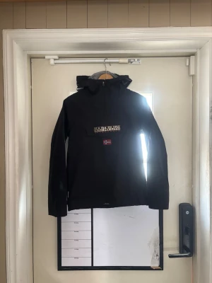 Svart vindjacka från Napapijri - Snygg svart vindjacka från Napapijri med huva och stor ficka framtill med logotyp och norsk flagga. Jackan har dragkedja vid halsen och är tillverkad i ett slitstarkt material som står emot vind. Perfekt för dig som vill ha en stilren och funktionell jacka. Storlek 162. Använd två gånger.