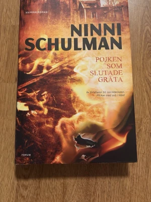 Pojken som slutade gråta - En spännande kriminalroman av Ninni Schulman. Omslaget har eldiga toner i orange, gult och svart med en dramatisk bild av eld och ett hus i bakgrunden. Boken är i ett hårt skal och har en modern, stilren design. Perfekt för dig som gillar deckare och mysterier.