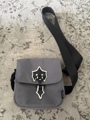 Grå axelväska med trapstar spänne - Snygg grå axelväska med justerbar svart rem och unik dekorativ spänndetalj i vitt och svart på framsidan. Väskan har lock och strukturmönstrad framsida, tillverkad i slitstarkt tygmaterial. Perfekt för dig som vill ha en cool och praktisk accessoar.