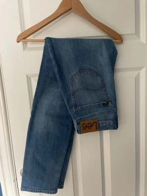 Blå raka jeans från Lee - Säljer ett par klassiska blå jeans från Lee med raka ben och hög midja. Jeansen har en stor bakficka och en brun läderpatch med Lee-loggan bak. Tillverkade i slitstark denim, perfekta för en avslappnad och trendig look. Storlek 29,32
