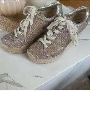 Glittriga kilklackade sneakers - Säljer ett par unika sneakers med kilklack och glittrigt tyg i beige och silver. Skorna har snörning, metallic-detaljer vid hälen och flätad sula i espadrillestil. Perfekta för dig som vill sticka ut med både höjd och bling.