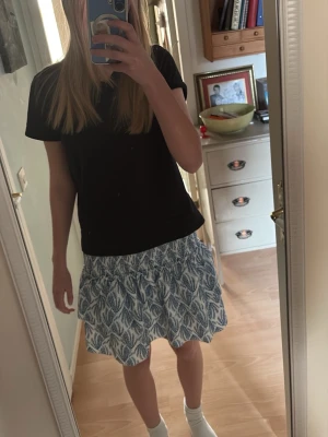 Blåvit kjol - Kort kjol med blått och vitt bladmönster. Kjolen har resår i midjan och är gjord i ett lätt, luftigt material som passar perfekt till varmare dagar. Snygg att matcha med en enkel topp för en avslappnad look.