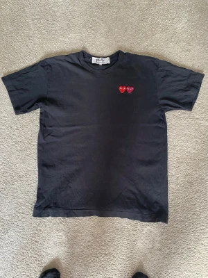 Svart Comme des Garçons PLAY t-shirt - Svart t-shirt från PLAY med två röda hjärtan med ögon broderade på bröstet. Klassisk passform och rund halsringning. Perfekt för dig som gillar stilrena plagg med en twist.