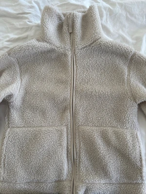 Beige teddyjacka med dragkedja - Mysig beige teddyjacka med hög krage och hel dragkedja framtill. Jackan har två stora fickor. Enkel att matcha med olika outfits. Jag är inte förtjust i just denna då jag inte tycker den passar min kroppsform därmed är den mycket sällan använd! 