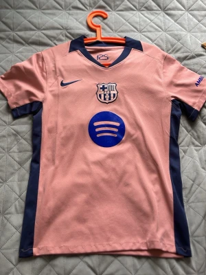 Rosa FC Barcelona matchtröja Nike - Säljer en rosa FC Barcelona matchtröja från Nike med klubbmärke och Spotify-logga på bröstet. Tröjan har mörkblå detaljer längs sidorna och vid kragen. Perfekt för dig som älskar fotboll och vill sticka ut på planen.