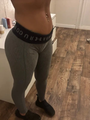 Grå leggings från Under Armour - Snygga grå leggings från Under Armour med bred svart midjeresår där loggan syns tydligt. De är tighta och stretchiga, perfekta för träning eller chill. Materialet känns mjukt och följsamt mot kroppen.