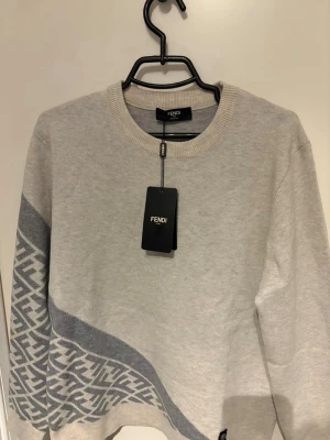 Beige stickad tröja från Fendi - Snygg beige stickad tröja från Fendi med rund halsringning och ett grått diagonalt mönster med Fendi-logga längs nederdelen och ena ärmen. Tröjan har ribbade muddar och är perfekt för dig som gillar exklusiv och trendig design. Endast testad på taggen är av med medföljer i paketet