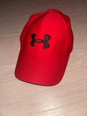 Röd keps från Under Armour - Snygg röd keps från Under Armour med klassisk böjd skärm och stort svart UA-logotyp broderat framtill. Modellen är UA Classic Fit och kepsen har paneler med sömmar för en sportig look. Perfekt för dig som gillar streetwear och sportig stil.