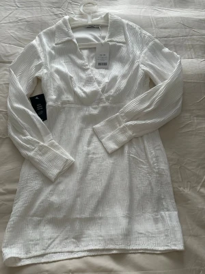 Vit kortklänning från NA-KD - Stilren vit kortklänning från NA-KD med långärmad design och subtilt randigt strukturmönster. Klänningen har krage, v-ringning och markerad midja för en snygg siluett. Perfekt för dig som gillar minimalistisk och trendig stil.