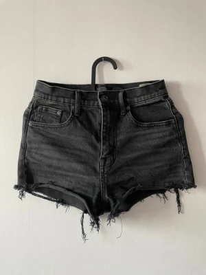 Crocker jeansshorts - Svarta högmidjade Crocker jeansshorts med råa, fransiga kanter och klassisk femficksdesign. Shortsen har knapp- och dragkedjestängning och är perfekta för en avslappnad stil. 