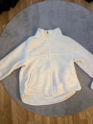 Vit teddyjacka med dragkedja - Mysig vit teddyjacka med hög krage och hel dragkedja framtill. Jackan har lång ärm och är tillverkad i fluffigt, mjukt material som ger en avslappnad och trendig look. Perfekt för kyliga dagar när du vill vara både varm och snygg. Använd några gånger, jätte mysig och skön! Inte fått användning till på senaste.