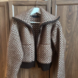 Brun och vit stickad bomberjacka - Säljer en snygg stickad bomberjacka från Zara i brunt och vitt rutmönster. Jackan har bred krage, dragkedja framtill och ribbade muddar. Perfekt för dig som vill ha en unik och mysig jacka med cool retrovibe.