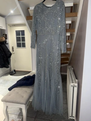 Ljusblå långklänning med paljetter - Säljer en elegant ljusblå långklänning med glittriga paljettdetaljer och långärmad design. Klänningen har rund halsringning och är tillverkad i ett tunt, skirt material som ger ett exklusivt intryck. Perfekt för dig som vill sticka ut med en glamorös look. Använd endast en gång.