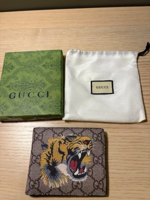 Gucci plånbok med tigertryck - Säljer en snygg brun plånbok från Gucci med klassiskt GG-mönster och ett färgstarkt tigertryck på framsidan. Plånboken är i canvasmaterial och har en fyrkantig form. Kommer med original ask och dustbag.(wallet)(005)