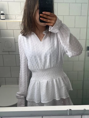 Vit volangklänning med smock i midjan - Superfin vit klänning med volangdetaljer och smockad midja. Klänningen har långärmade ärmar, v-ringning och ett diskret prickigt mönster i tyget. Perfekt för sommarens alla tillfällen och ger en romantisk vibe. säljer den då för den inte kommer till användning, skriv om ni har frågor 💕