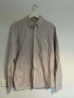 Randig skjorta från John Henric  - Klassisk långärmad skjorta med smala vertikala ränder i vitt och ljusrosa . Skjortan har knappar framtill och på ärmsluten samt en stilren krage. Tillverkad i mjuk bomull som känns skön mot huden. Perfekt för dig som gillar en clean och tidlös look. Funkar perfekt till ett uppklädd tillfälle men även till en lite mer avslappnad look. 